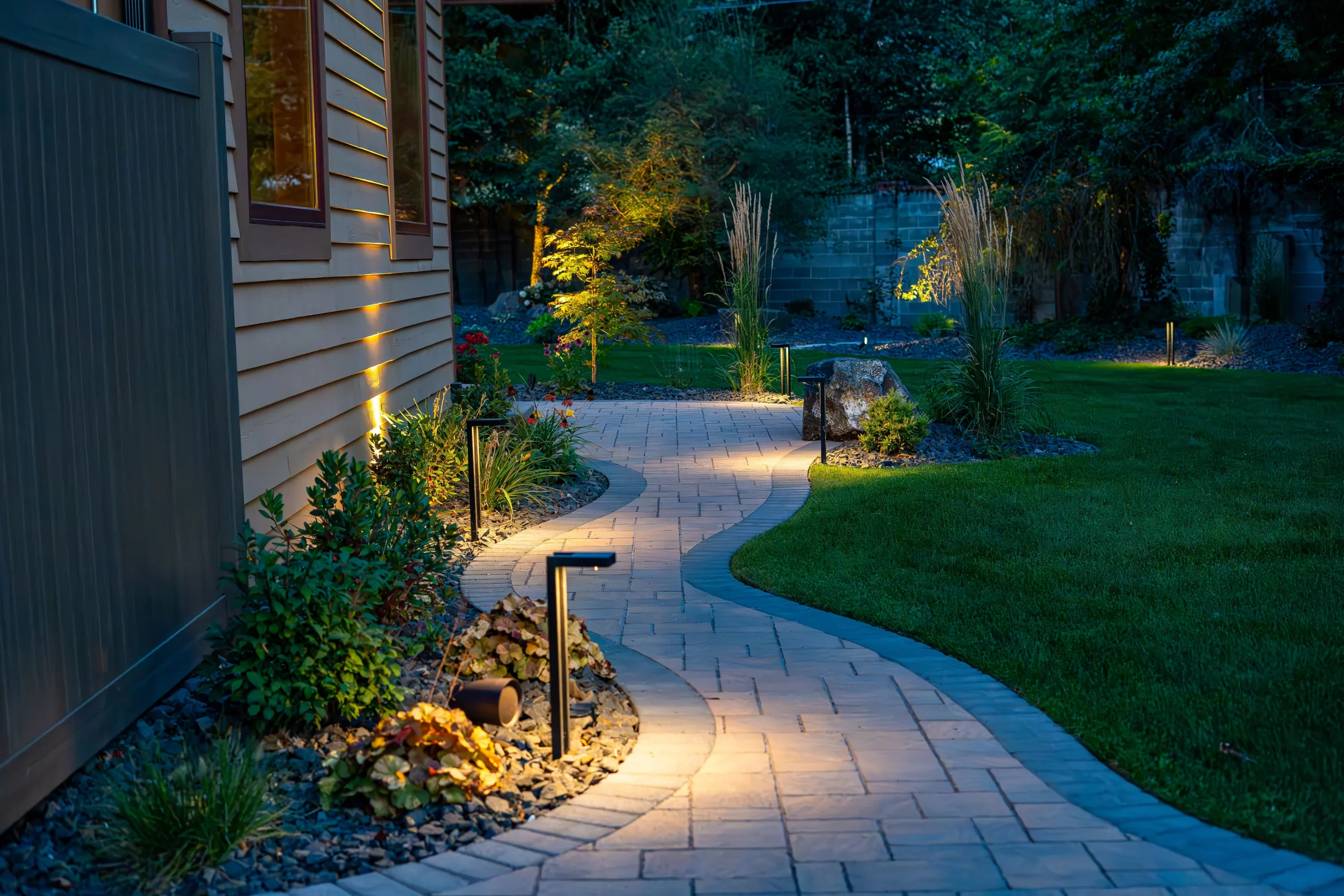 Representative Legacy Landscapes portfolio: curving paved residential walkway with lawn, planting beds, trees, and low landscape lighting.