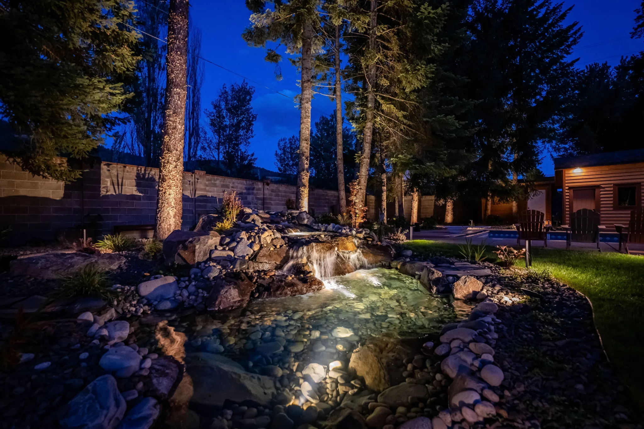 Representative Legacy Landscapes portfolio: illuminated natural-rock stream and waterfall in a wooded residential landscape.